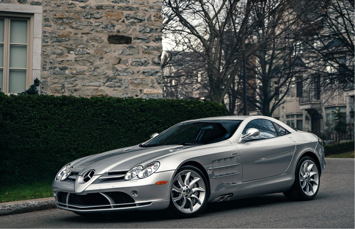 A silver Mercedes-Benz SL Rodeo sports car parked on a street in front of a stone house and bare trees.